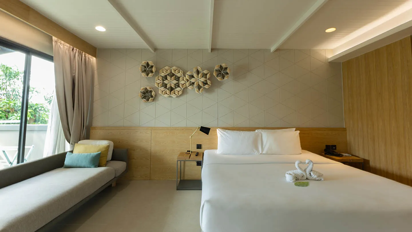 A large bed with swan towels, wooden bedside tables with lamps, a sofa with cushions, and light wood décor accented by a floral-patterned wall art for a serene, airy ambiance