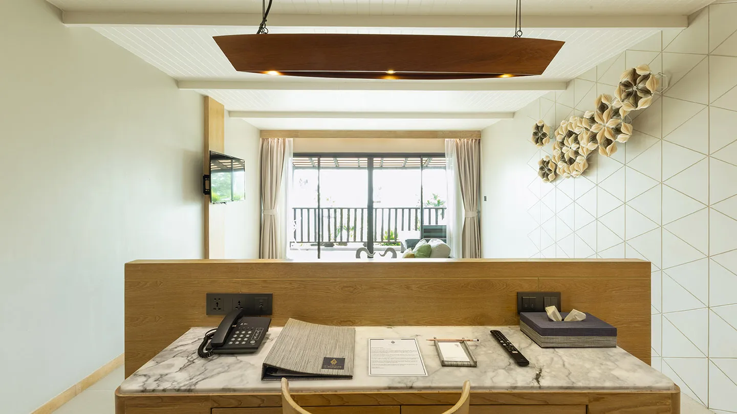 The Canna Deluxe room features a desk with a marble-patterned top, modern Thai-inspired décor, and large windows opening to a balcony with views of greenery and the Kata Noi Beach