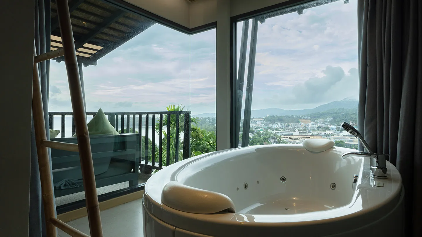 A spacious round bathtub faces floor-to-ceiling windows, showcasing Kata Noi Beach from a modern, warmly lit room with wooden touches