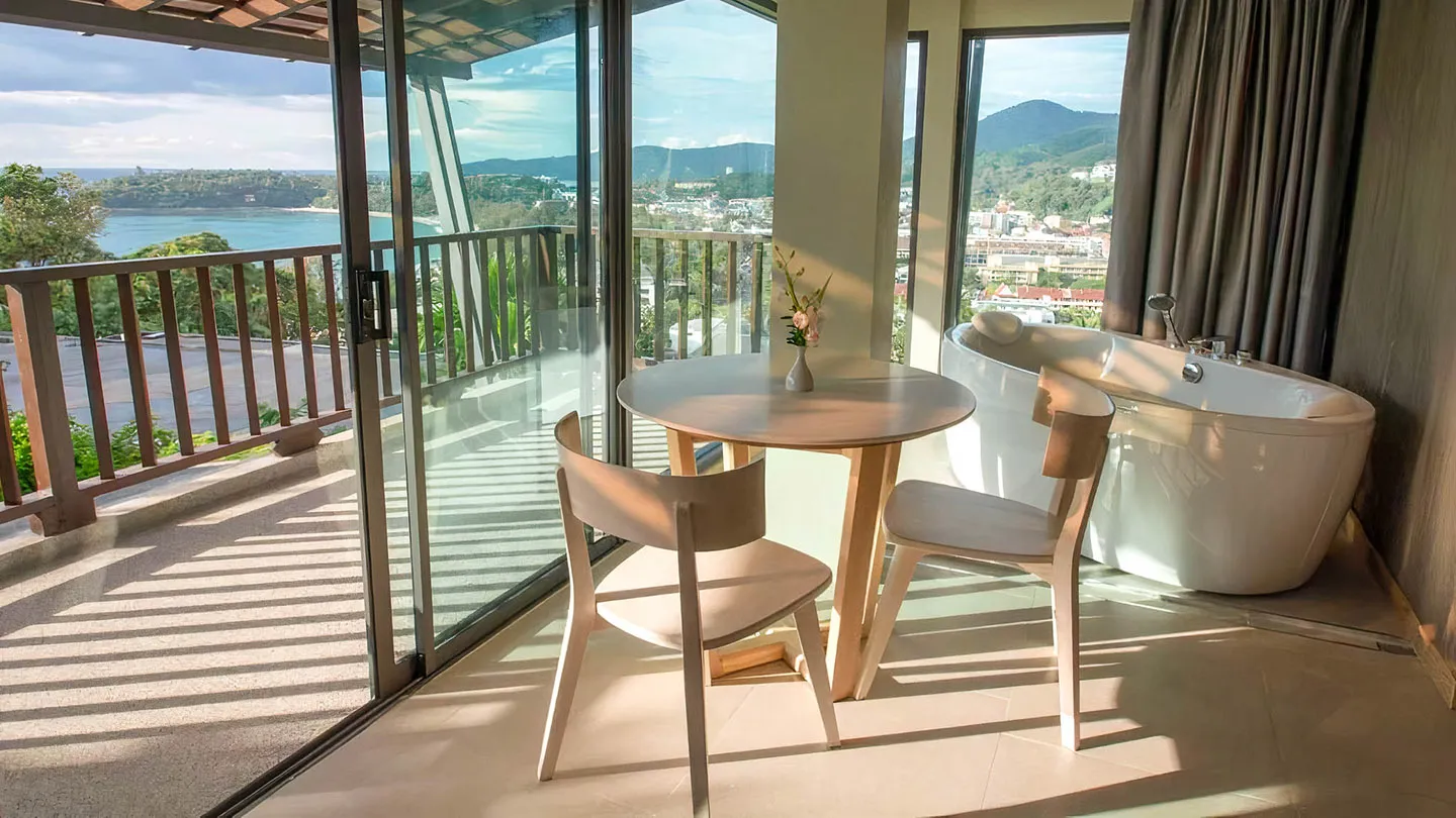 The suite’s private balcony overlooks Kata Beach, featuring an outdoor bathtub and an indoor seating area with a table and two chairs for a serene, romantic retreat