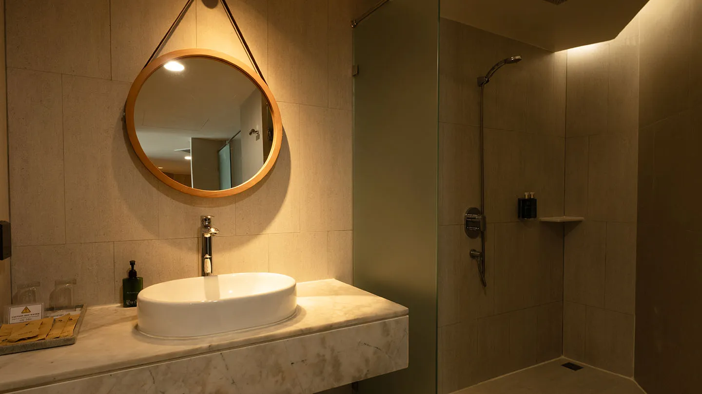 A bright Junior Suite bathroom featuring a vessel sink, round mirror, soft lighting, and a glass-enclosed rain shower with a clean, natural aesthetic