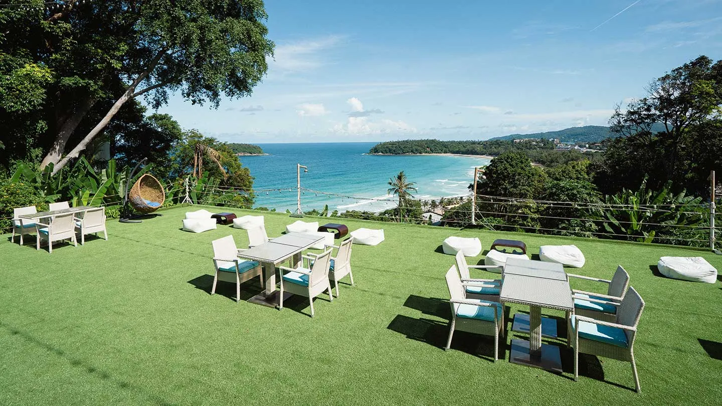 A panoramic outdoor scene of a lush resort lawn with pathways, shaded seating areas, swinging basket chairs, a glimpse of a pool, and a breathtaking view of the Kata Noi Beach