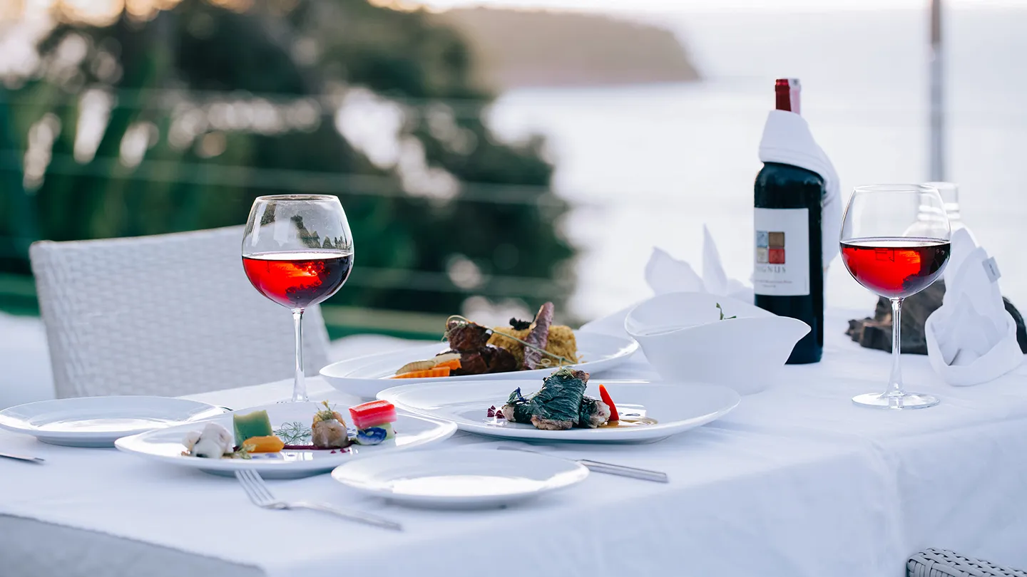 The table is set with multiple courses of food and two glasses of red wine, along with a bottle of wine wrapped in a white cloth. The setting appears to be outdoors, with a scenic view of the Andaman Sea and Kata Beach