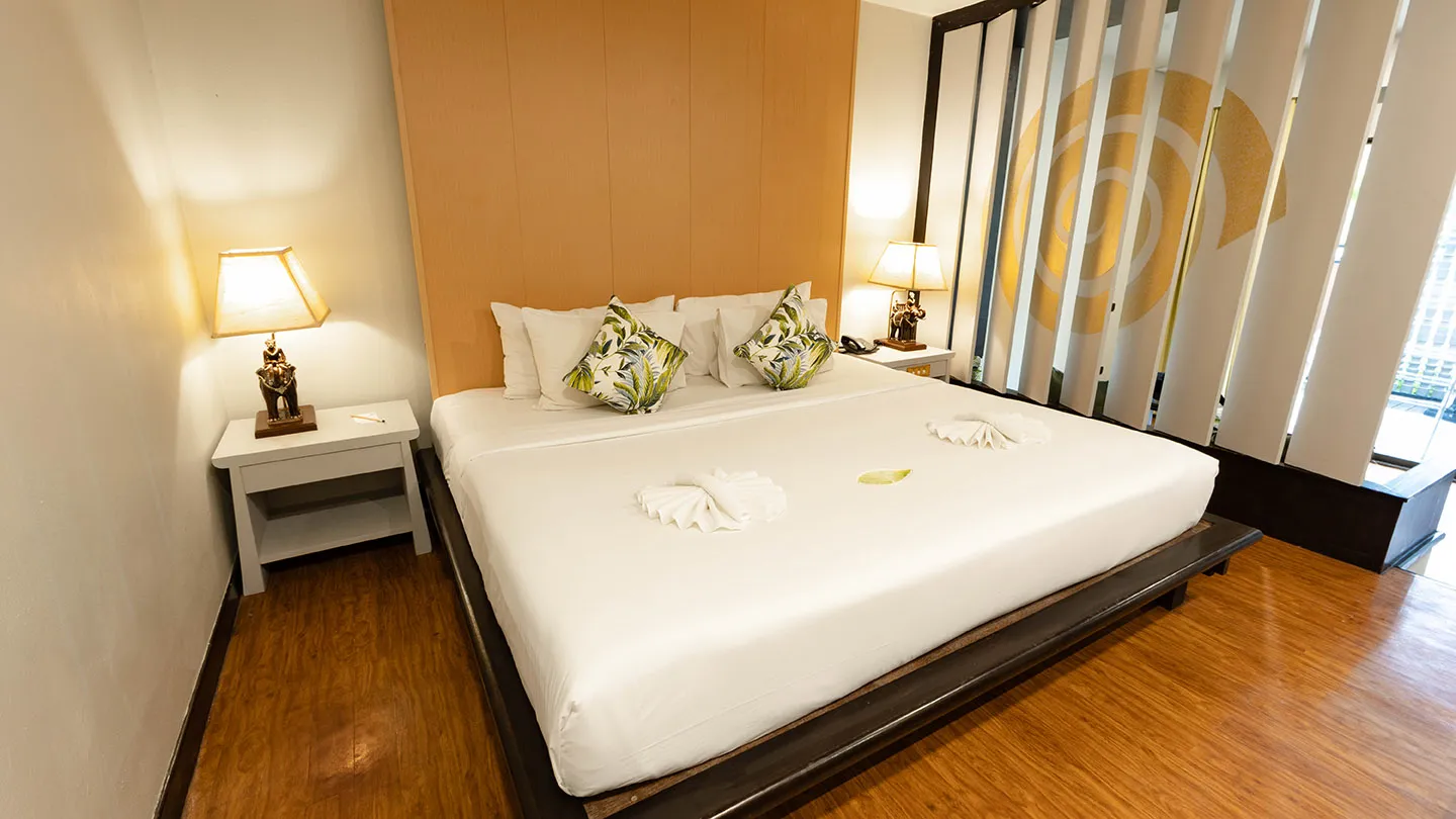 A spacious bedroom featuring a neatly made king-size bed with decorative pillows and rolled towels, bedside lamp and phone, light wood-paneled walls, dark flooring, and a window with sheer curtains