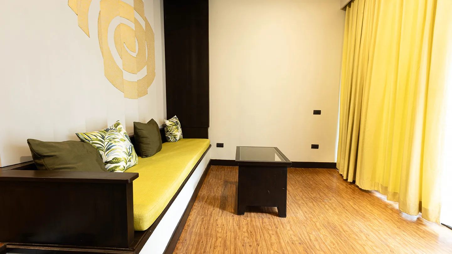 A long bench-style sofa with yellow cushions and pillows, a dark coffee table, wood-style flooring, white walls featuring a large rose mural, and yellow curtains