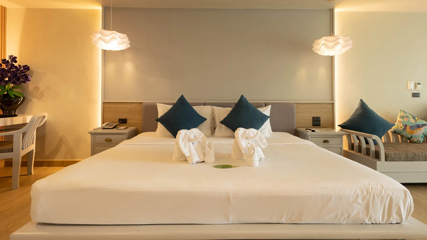 A spacious, light-filled room featuring a white bed with elephant towel decor, chic lamps, a blue-accented headboard, and a cozy seating area in modern Thai style