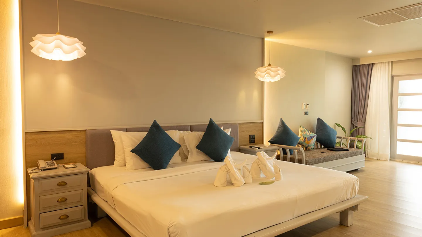 A stylish, well-furnished bedroom featuring a large bed, side tables with lamps, a seating area, and modern Thai-inspired decor in soft, natural tones