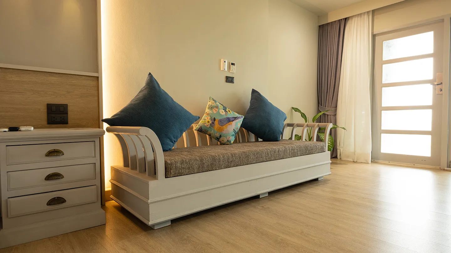 A sunlit, tranquil room with wooden floors, light walls, a blue-cushioned daybed, wooden dresser, and glass doors leading to lush greenery