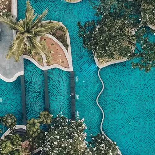 The resort showcases a cascading double-level pool amid lush greenery, sun loungers, and scenic sea views from above