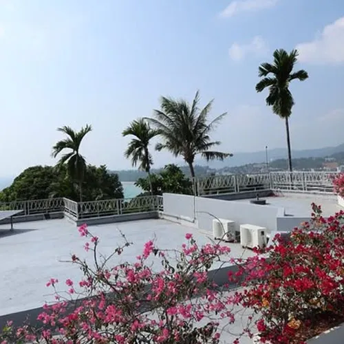 A scenic beach resort view of the Andaman Sea, framed by vibrant flowers, lush greenery, palm trees, and a light-colored building with a terrace