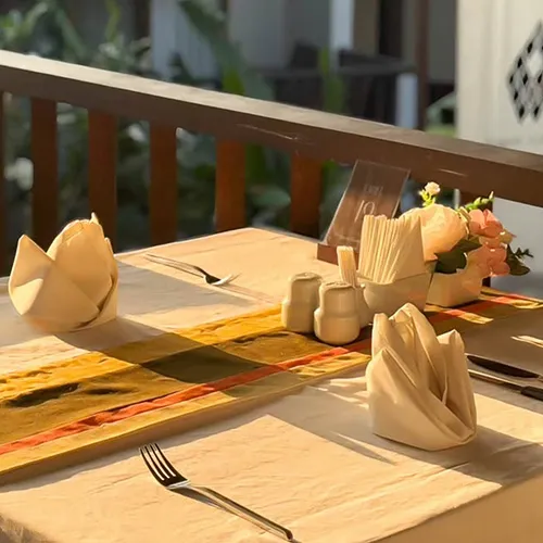 A table with an earthy-toned runner is set with plates, utensils, folded napkins, and a centerpiece of condiments and a small floral arrangement